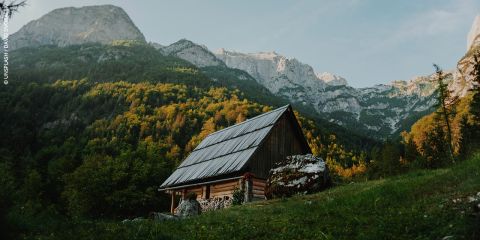 Eine Holzhütte mit schrägem Dach steht an einem grasbewachsenen Hang, umgeben von bewaldeten Bergen unter einem teilweise bewölkten Himmel.