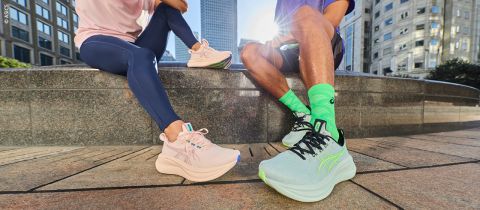 Zwei Personen in Sportkleidung sitzen im Freien auf einem Steinsockel und zeigen pastellfarbene ASICS-Laufschuhe, während im Hintergrund städtische Gebäude zu sehen sind.