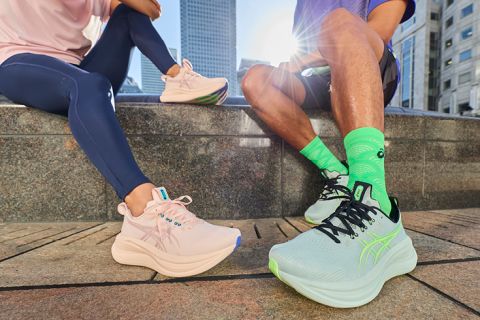 Zwei Personen in Sportkleidung sitzen im Freien auf einem Steinsockel und zeigen pastellfarbene ASICS-Laufschuhe, während im Hintergrund städtische Gebäude zu sehen sind.