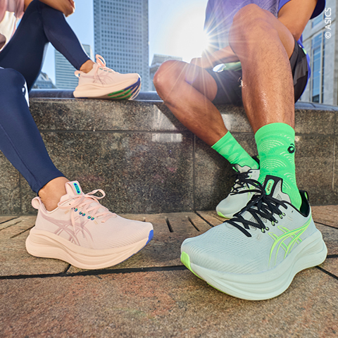 Zwei Personen sitzen im Freien und tragen ASICS-Laufschuhe - ein Paar ist hellrosa, das andere blassblau mit neongrünen Akzenten und passenden Socken.