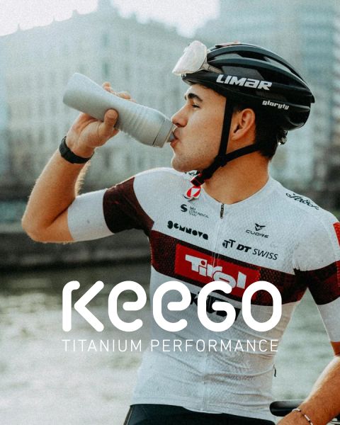 Ein Radfahrer in einem weiß-roten Trikot trinkt aus einer Flasche und trägt einen Helm, auf dem der Text KEEGO Titanium Performance" eingeblendet ist.