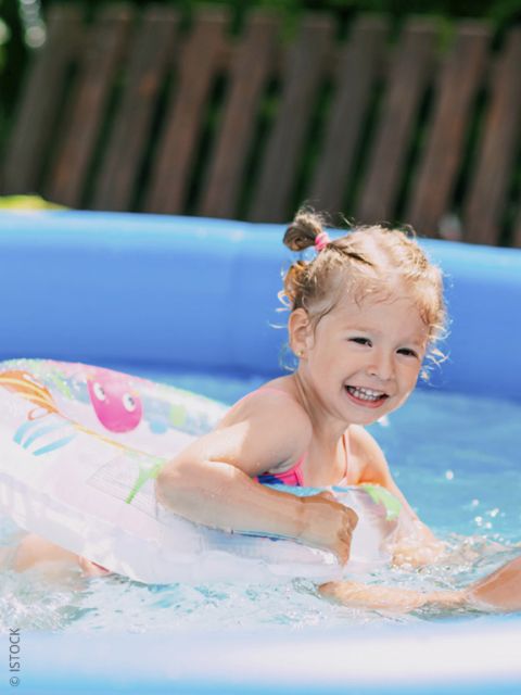 Ein lächelndes junges Mädchen mit einem Schwimmflügel planscht in einem kleinen blauen Pool im Freien an einem sonnigen Tag.