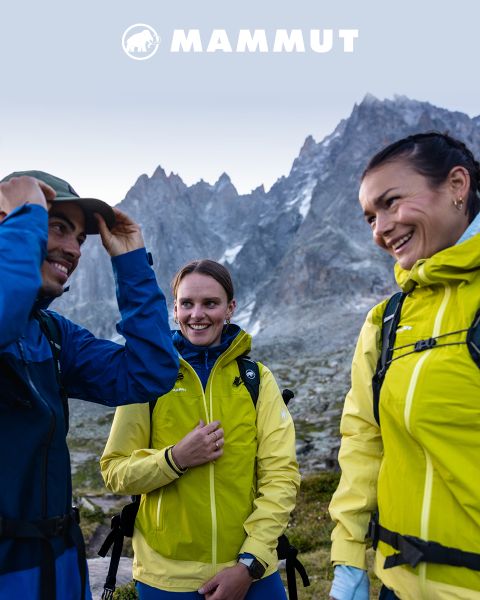 Drei Personen in hellen Outdoor-Jacken lächeln sich in einer Berglandschaft an, über ihnen das Mammut-Logo vor einem klaren Himmel.