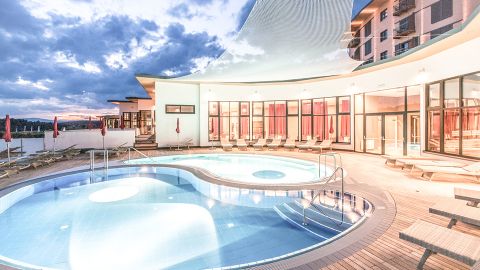 Moderner Außenpoolbereich eines Resorts mit Liegestühlen, geschwungener Architektur, großen Fenstern und Abendhimmel im Hintergrund.