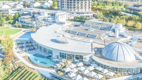 Luftaufnahme eines modernen Hotelkomplexes mit einer Kuppel, glasüberdachten Gebäuden, Außenpool, Gärten und Sonnenliegen an einem sonnigen Tag.