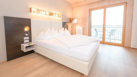 Helles, modernes Hotelzimmer mit Doppelbett, weißem Bettzeug, Holzfußboden, Wandleuchten und einem Balkon mit zwei Stühlen und einem Tisch.