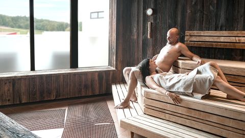 Ein Mann und eine Frau entspannen sich auf Holzbänken in einer Sauna; die Frau legt sich mit einem Handtuch hin, der Mann sitzt neben ihr.