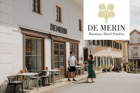 Ein händchenhaltendes Paar spaziert auf einer sonnigen Straße am DE MERIN Boutique Hotel Straden vorbei, an dessen Ecke das Logo des Hotels prangt.