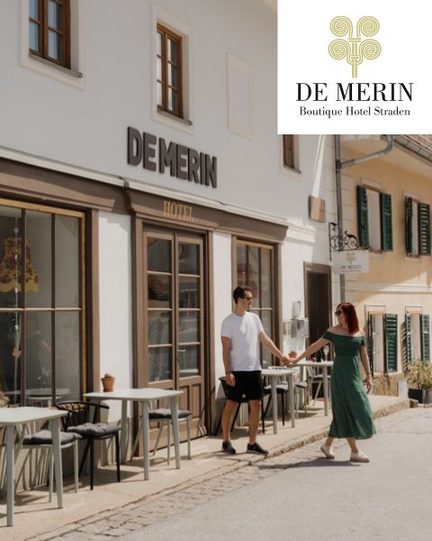 Ein Paar hält sich vor dem De Merin Boutique Hotel Straden mit Sitzgelegenheiten im Freien und einer sonnenbeschienenen Straße die Hand. Das Logo des Hotels erscheint in der oberen Ecke.
