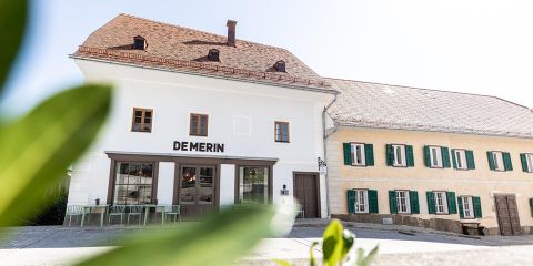 Straßenansicht eines weißen Gebäudes mit "DEMERIN"-Schild, Tischen im Freien und einem angrenzenden Haus mit grünen Fensterläden, gesehen durch unscharfe Blätter.