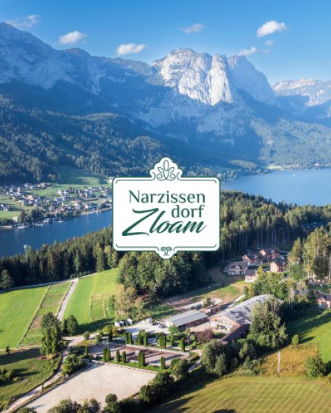 Ein malerisches Dorf an einem See und Bergen mit dem Logo "Narzissendorf Zloam" in der Mitte. Grüne Felder, Bäume und Häuser füllen den Vordergrund.