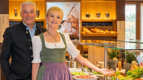 Ein lächelnder Mann und eine Frau in traditioneller Kleidung stehen an einem Buffet mit frischen Salaten und Brot in einem warm beleuchteten Restaurant.