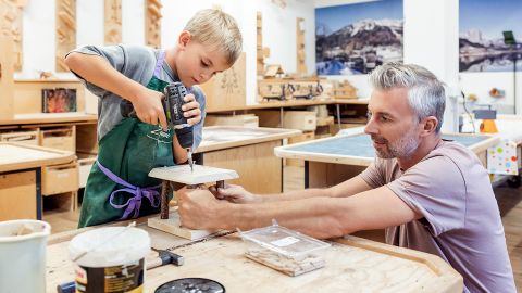 Ein kleiner Junge bohrt mit Hilfe eines erwachsenen Mannes in einer Werkstatt voller Werkzeuge und Holzbearbeitungsprojekte in Holz.