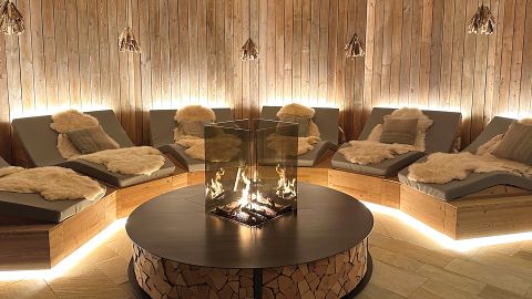 Gemütliche Lounge mit geschwungenen Holzwänden, gepolsterten Liegestühlen, Fellüberwürfen und einer modernen gläsernen Feuerstelle in der Mitte, die ein warmes Licht abgibt.