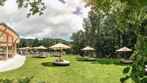 Lounge-Bereich im Freien mit runden, gepolsterten Liegen und Sonnenschirmen auf einer Rasenfläche, umgeben von Bäumen und einem modernen Glasgebäude auf der linken Seite.