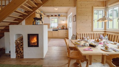 Gemütlicher hölzerner Essbereich mit Frühstücksgedeck auf dem Tisch, einem beleuchteten Kamin, gestapeltem Brennholz und Blick in die helle Küche.