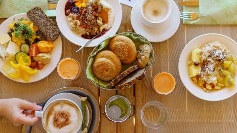 Ein Frühstückstisch mit Kaffee, Saft, Brötchen, Müslischalen mit Obst und Nüssen, Salat, Käse und Besteck auf einem Platzdeckchen.
