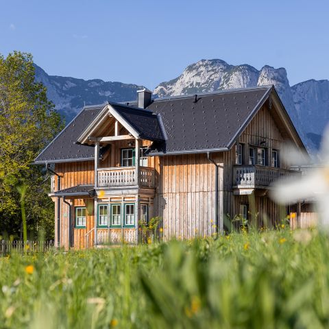Ein Holzhaus mit Balkon steht auf einer grasbewachsenen Wiese, mit Bergen und einem klaren blauen Himmel im Hintergrund.