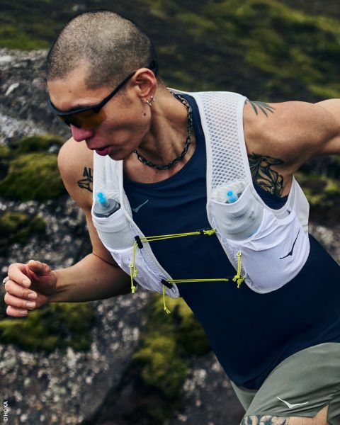 Eine Frau in Sportkleidung und mit Mütze trägt eine Laufweste mit Wasserflaschen und steht seitlich vor einem schlichten weißen Hintergrund.