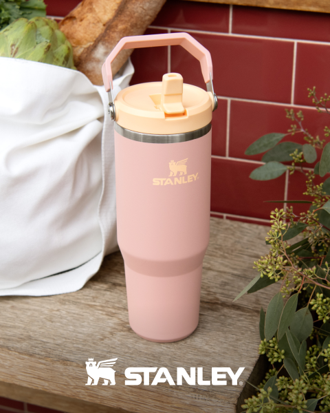 Ein rosafarbener Stanley-Becher mit Henkel steht auf einer Holzfläche neben einer weißen Tasche und grünen Pflanzen, mit dem Stanley-Logo am Boden.