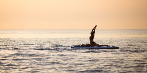 Eine Person praktiziert Yoga auf einem Paddelbrett im ruhigen Meerwasser bei Sonnenuntergang und streckt ihre Arme gegen den orangefarbenen Himmel in die Höhe.
