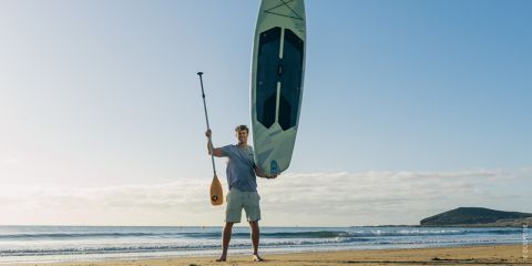 Ein Mann steht an einem Sandstrand, in der einen Hand ein Paddel, in der anderen ein aufrechtes Paddleboard, im Hintergrund das Meer und der Himmel.