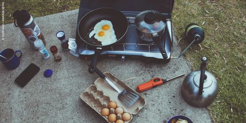 Eine Campingkochstelle mit Eiern, die auf einem tragbaren Herd gebraten werden, einem Topf, einem Kessel, Eiern, Gewürzen, einem Feuerzeug, Bechern und Utensilien auf Gras und Beton.