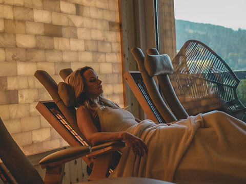 Eine Frau entspannt sich auf einem Liegestuhl, zugedeckt mit einer Decke, in einem schwach beleuchteten Wellness-Raum mit einem großen Fenster und einer Salzsteinwand.