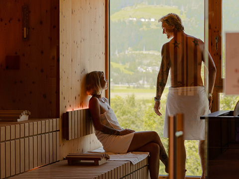 Zwei Personen entspannen sich in einer holzgetäfelten Sauna, eine sitzend und eine stehend an einem großen Fenster mit Blick ins Freie.