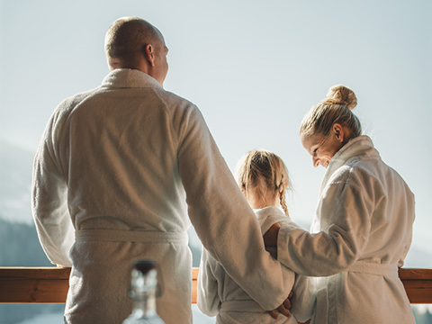 Eine Familie in weißen Gewändern steht auf einem Balkon und blickt auf eine malerische Landschaft, während das Sonnenlicht von hinten auf sie scheint.