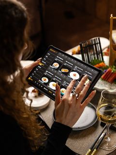 Eine Person blättert auf einem Tablet in einer digitalen Speisekarte an einem Restauranttisch, der mit Essen, Stäbchen und einem Glas Weißwein gedeckt ist.