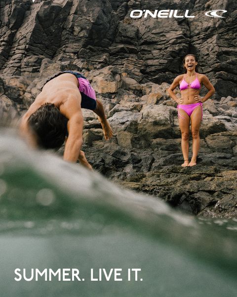 Ein Mann taucht in der Nähe von Felsen ins Wasser, während eine Frau in einem rosa Bikini lächelnd daneben steht. Der Text lautet: "SUMMER. LIVE IT." und das O'Neill-Logo ist zu sehen.