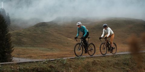 Zwei Radfahrer fahren auf einer asphaltierten Straße durch eine neblige, hügelige Landschaft, im Hintergrund sind Gras und Bäume zu sehen.
