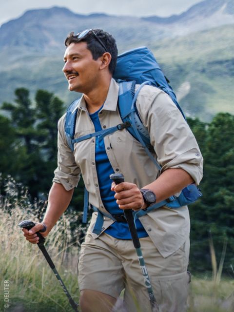 Ein Mann, der mit Wanderstöcken in der Natur wandert, einen Rucksack, ein beiges Hemd und Shorts trägt und vor dem Hintergrund grüner Bäume und Berge lächelt.