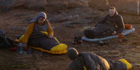 Drei Menschen in Schlafsäcken sitzen bei Sonnenaufgang im Freien auf felsigem Untergrund, bereiten Essen zu und lachen gemeinsam beim Campen.
