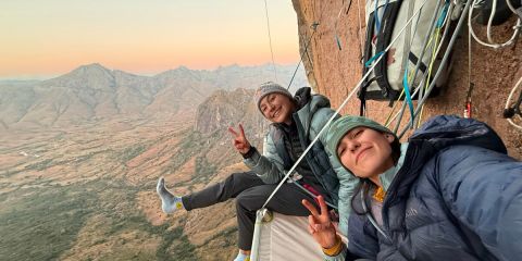 Zwei Personen in Kletterausrüstung lächeln und zeigen das Peace-Zeichen, während sie auf einer Portaledge hoch oben an einer Felswand ausruhen, unter ihnen erstrecken sich Berge und die Landschaft.