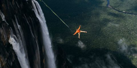 Eine Person in einem orangefarbenen Anzug macht einen Bungee-Sprung in der Nähe eines hohen Wasserfalls, umgeben von üppigem, grünem Wald und einem sich schlängelnden Fluss unterhalb.