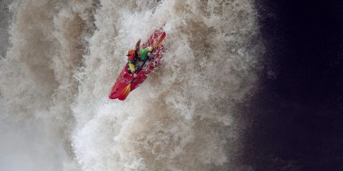 Ein Kajakfahrer, der einen Helm und Schutzkleidung trägt, stürzt sich in einen gewaltigen, reißenden Wasserfall, umgeben von tosendem Wildwasser.