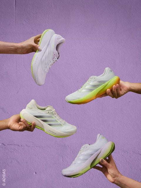Vier Hände, die weiße Sportschuhe mit grünen und gelben Sohlen vor einer lavendelfarbenen Wand halten.