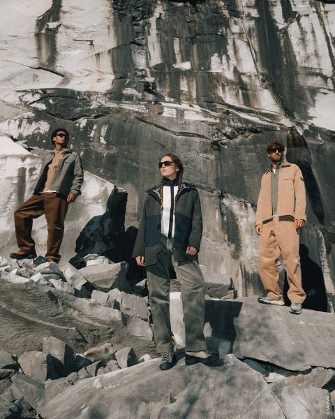 Drei Personen in Outdoor-Kleidung stehen auf felsigem Gelände, hinter ihnen eine große Steinmauer, und blicken bei strahlendem Sonnenschein in die Ferne.