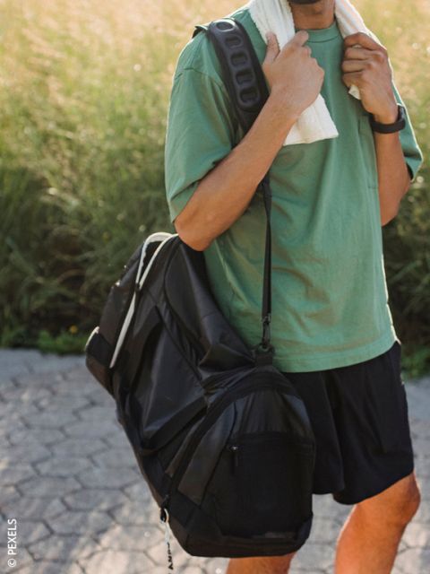 Eine Person in einem grünen Hemd, die ein Handtuch um den Hals trägt, trägt an einem sonnigen Tag draußen eine schwarze Sporttasche.
