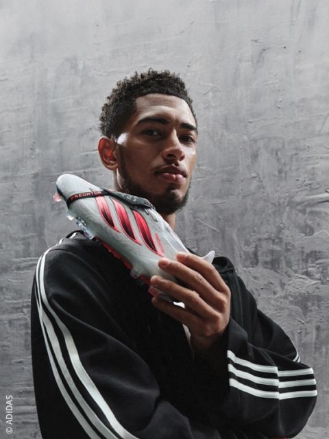 Ein Mann in einem Adidas-Trainingsanzug hält einen grau-roten Adidas-Fußballschuh vor sein Gesicht und steht vor einem strukturierten grauen Hintergrund.