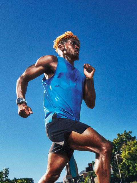 Ein Mann in einem blauen Tanktop und schwarzen Shorts läuft an einem sonnigen Tag im Freien, im Hintergrund sind Gebäude und Bäume zu sehen.