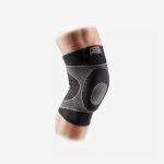 Eine Person, die eine schwarz-graue Kniebandage am angewinkelten Bein trägt, vor einem schlichten weißen Hintergrund.