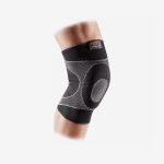 Eine Person, die eine schwarz-graue Kniebandage am angewinkelten Bein trägt, vor einem schlichten weißen Hintergrund.