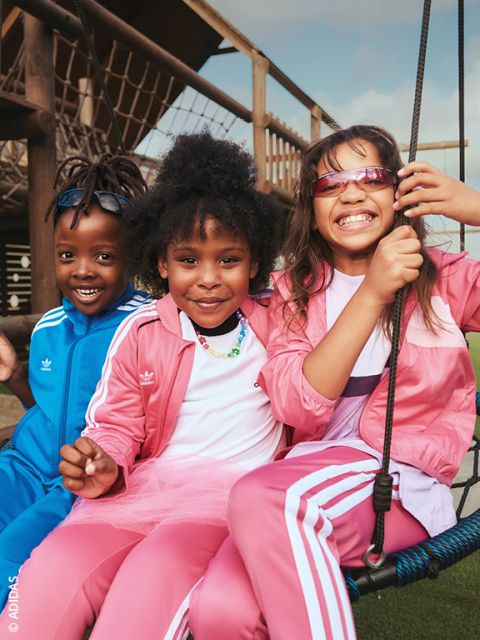 Drei lächelnde Kinder in Adidas-Outfits sitzen draußen auf einer Schaukel – zwei davon in rosa Trainingsanzügen und eines in einem blauen – und genießen den sonnigen Tag.