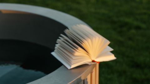 Ein aufgeschlagenes Buch, dessen Seiten aufgefächert sind, liegt am Rand einer runden Metallfläche in der Nähe einer Rasenfläche.