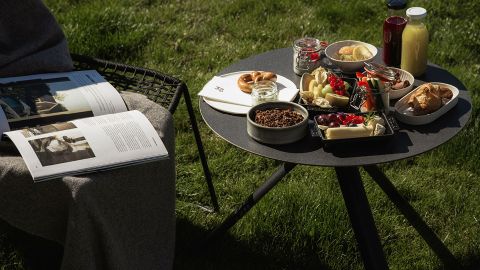 Ein runder Tisch auf dem Rasen mit verschiedenen Frühstücksspeisen und Getränken, daneben eine Person, die eine Zeitschrift liest und teilweise mit einer Decke zugedeckt ist.