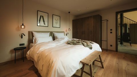 Ein gemütliches Schlafzimmer mit einem Doppelbett, weißer Bettwäsche, zwei Hängeleuchten, Wandbildern, einer Holzbank, einem Kleiderschrank und einer offenen Tür zu einem anderen Raum.