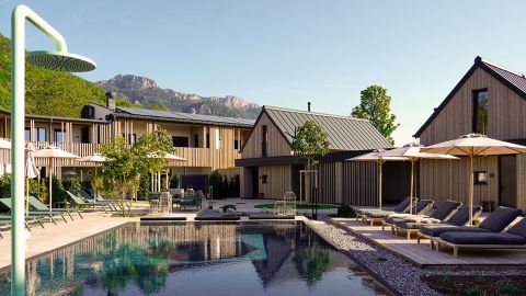 Moderne Holzgebäude umgeben einen Pool mit Liegestühlen und Sonnenschirmen, im Hintergrund bietet sich unter strahlend blauem Himmel ein Blick auf die Berge.
